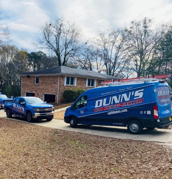Two blue Dunn’s HVAC, plumbing, and electrical service vehicles are parked in the driveway of a two-story brick suburban house on a clear day.