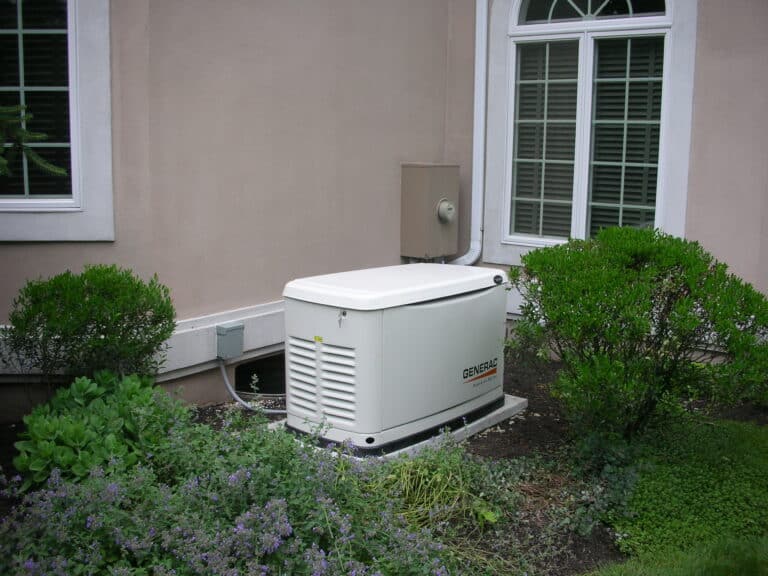A standby generator is installed outside a house, positioned near bushes and a window with white shutters.