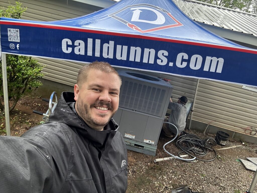 Man in rain jacket smiling in front of HVAC unit while a technician works under a "calldunns.com" canopy outside a Birmingham home.