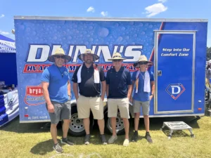 Four people wearing hats and sunglasses stand in front of a Dunn’s HVAC trailer on a sunny day, with towels around their necks.