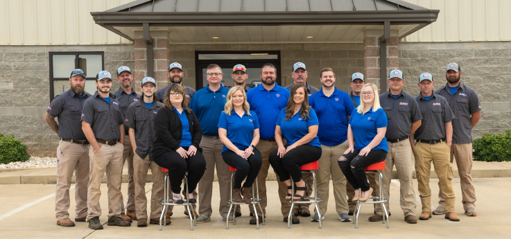 A group of eighteen people, some seated and some standing, pose outside a building; most are wearing blue shirts or matching uniforms.
