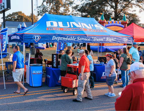 People visit a Dunn's HVAC, Plumbing, and Electrical booth at an outdoor event, with banners, staff, and a bouncy house visible in the background under sunny weather.