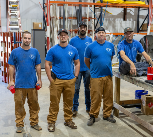 Five men in blue shirts and brown pants stand and sit in a workshop with shelves, ladders, and equipment in the background.
