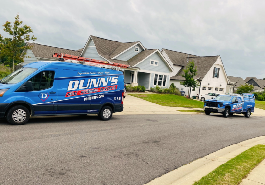 Two blue Dunn’s Heating, Cooling & Plumbing service vehicles are parked on a residential street in front of modern suburban houses.