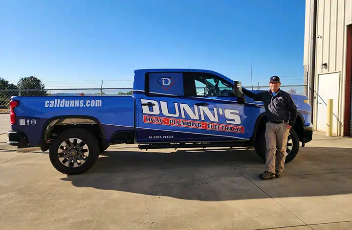 A person stands next to a blue Dunn's service truck labeled “HVAC, Commercial Plumbing, Electrical” parked outside a building on a sunny day.