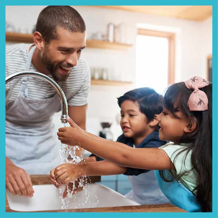 A smiling man helping two children wash their hands at a kitchen sink.