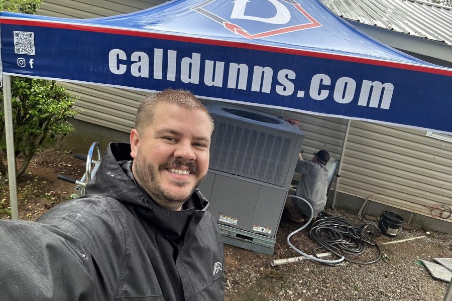 Man in rain jacket smiling in front of HVAC unit while a technician works under a "calldunns.com" canopy outside a Birmingham home.